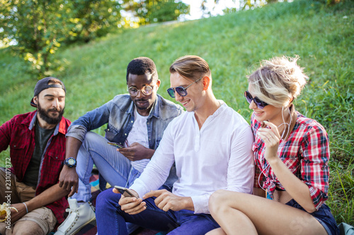 Multiracial group of carefree people dressed in casual summer wear sitting on park lown and watching something intresting in smartphone. Women and men wearing casual stylish clothes and sunglasses. © alfa27