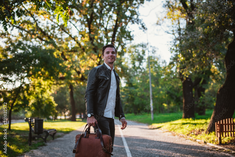 Young handsome man with bag walking in park during sunny day, autumn theme