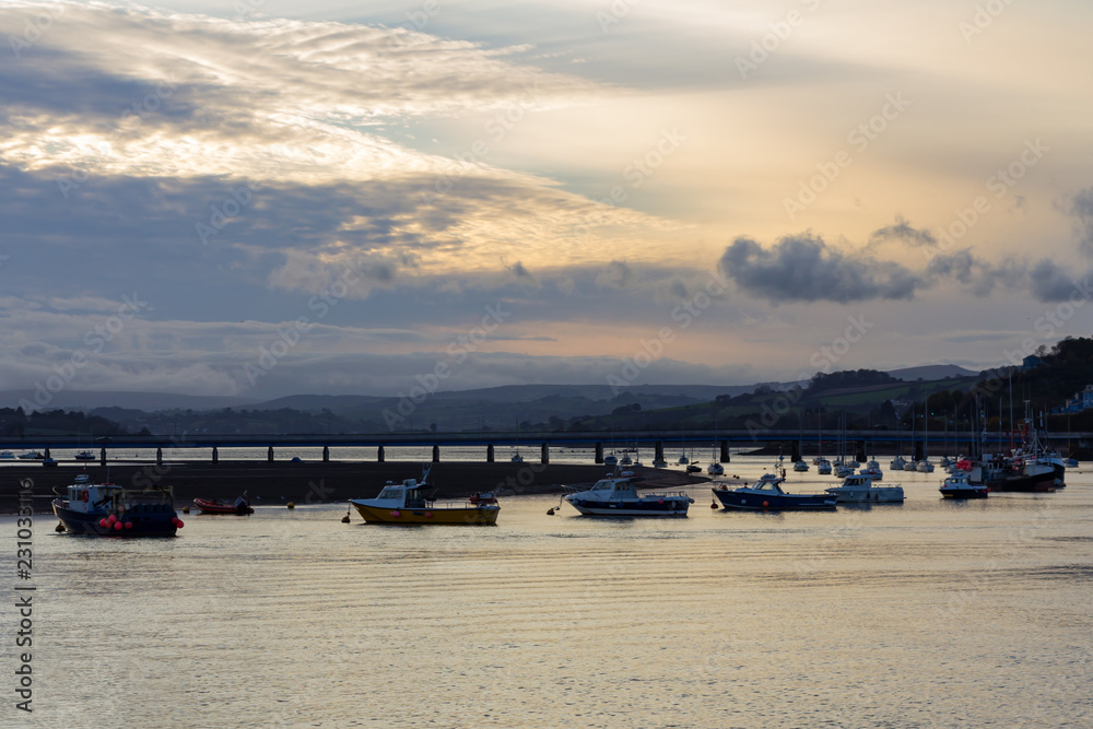 Fishing boats moored in the River Teign estuary at sunset under a cloudy sky with the Shaldon bridge in the background