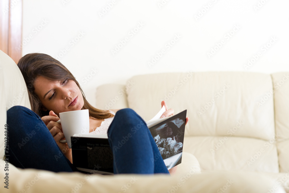 Brunette and young woman quietly reading a book while drinking hot coffee, tea or infusion cup stretched on the couch in the living room of her house Brunette and young woman quietly reading a book while drinking hot coffee, tea or infusion cup stretched on the couch in the living room of her house