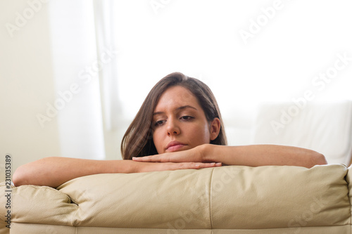 Serious brunette girl leaning on her two arms on a beige sofa looking to the side thoughtfully with her hair behind her ear