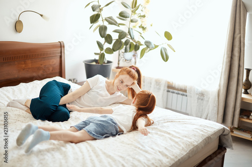 Caucasian ginger haired woman and her red-haired daughter wearing home clothing enjoying spare time in the morning, lying in bed at home and talking about wishes and dreams. photo