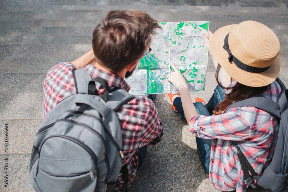Man and woman sits on steps and holds map together. They look at it. Tourists have rocksacks on back. Woman wears hat. Man and woman sits on steps and holds map together. They look at it. Tourists have rocksacks on back. Woman wears hat.