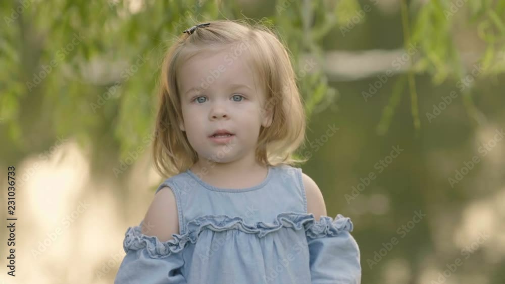 Portrait of little girl looks in cam and smiling under the tree near the lake in the sunset light. Trees and sunset reflected in water. Baby emotions - happy laugh. Portrait of little girl looks in cam and smiling under the tree near the lake in the sunset light. Trees and sunset reflected in water. Baby emotions - happy laugh.