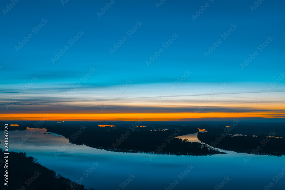 Beautiful river with big island with trees under predawn sky. Bright orange stripe in picturesque cloudy sky. Early blue sky reflected in water. Colorful morning atmospheric image of majestic nature.