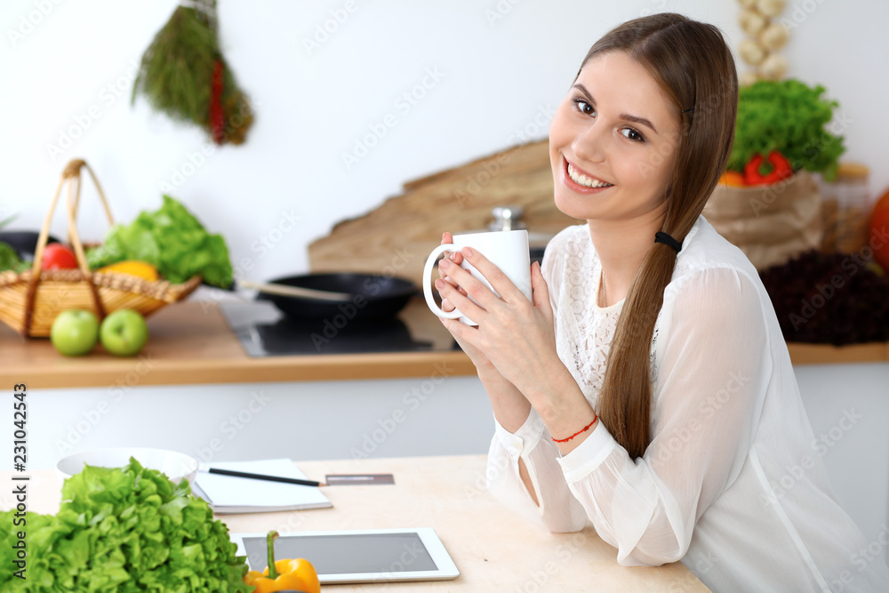 Young happy woman holding white cup and looking at the camera while sitting at wooden table in the kitchen among green vegetables. Good morning, lifestyle or cooking concept Young happy woman holding white cup and looking at the camera while sitting at wooden table in the kitchen among green vegetables. Good morning, lifestyle or cooking concept