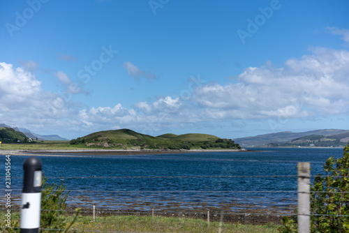 Sicht von der Stra  e zwischen zwei Pfosten sieht man einen Zaun Weide und dahinter ein See   ein paar wei  en Wolken am blauen Himmel Schottland