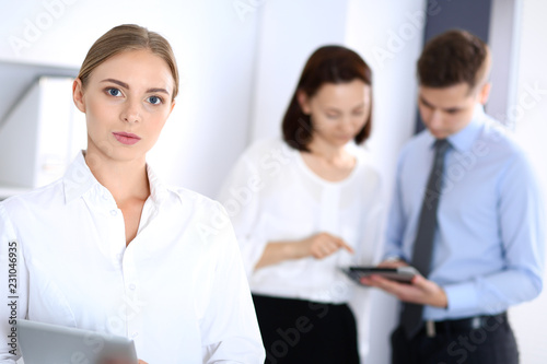 Beautiful blonde businesswoman standing straight in a brightly lit office at the background of colleagues or partners. Business concept