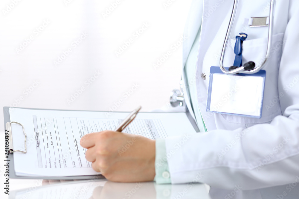 Female doctor filling up an application form with silver pen while sitting at the desk in hospital office. Just hands. Medicine and health care concept Female doctor filling up an application form with silver pen while sitting at the desk in hospital office. Just hands. Medicine and health care concept