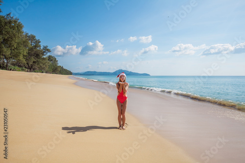 Happy young woman with long hair in red swimsuit and santa claus hat sending air kiss while standing on the tropical beach by the sea with beautiful blue water and blue sky on Phuket island,Thailand