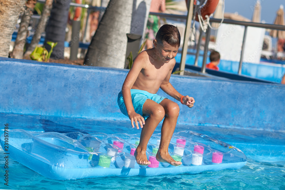 Naklejka premium Smiling Caucasian boy having fun in swimming pool at resort. He is standing on inflatable mattress and ready to jump from it.