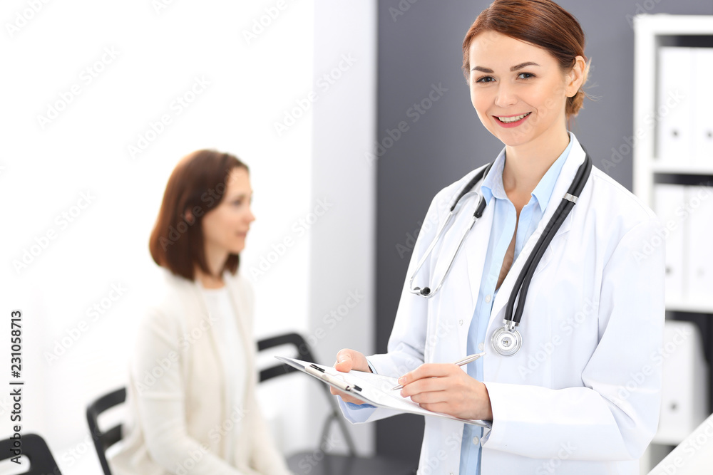 Doctor woman at work. Portrait of female physician filling up medical form while standing near reception desk at clinic or emergency hospital. Patient woman sitting at the background. Medicine concept Doctor woman at work. Portrait of female physician filling up medical form while standing near reception desk at clinic or emergency hospital. Patient woman sitting at the background. Medicine concept