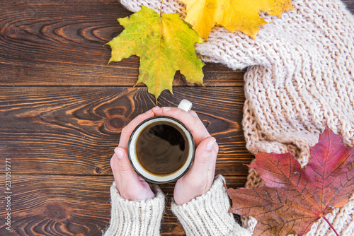 Hand holding mug of coffee on dark wooden background with autumn leaves and scarf