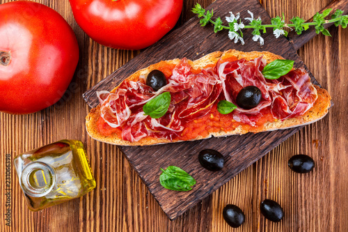 Delicious bread toast with natural tomato, extra virgin olive oil, Iberian ham, black olives and basil leaves. On wooden background.