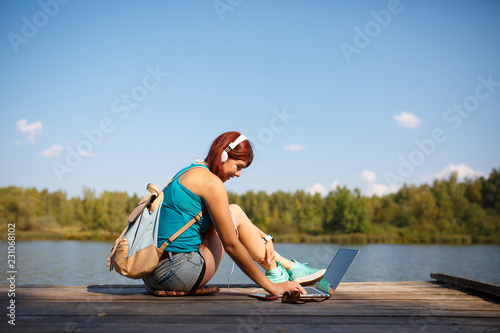 Image of woman with headphones in headphones with backpack and laptop in hands sitting on bank of river