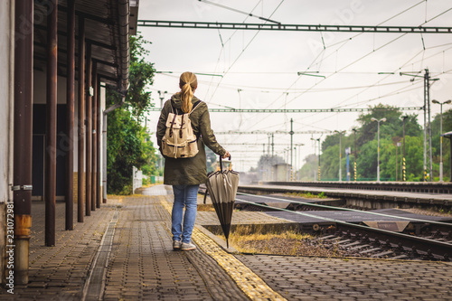Woman with backpack and umbrella is waiting for a train on railroad station. Travelling during rain at autumn season. Travel concept 