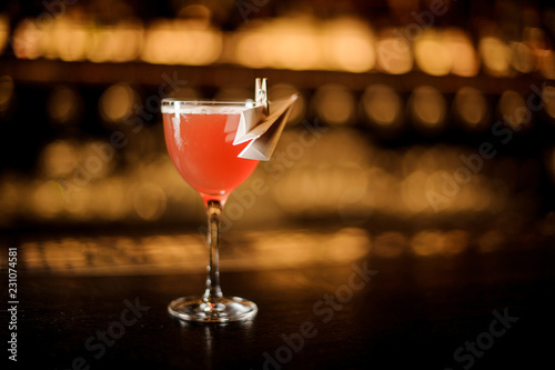Glass of a delicious red cocktail decorated with a paper plane standing on the bar counter