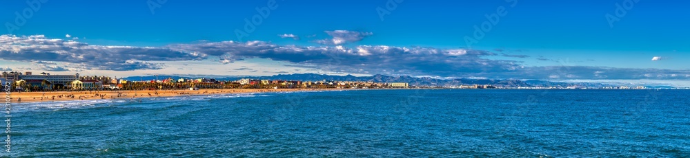 A bright sunny panoramic view of the blue waters off Cabanyal beach in Valencia Spain with the buildings and people dotting the shoreline and mountains in the distance.
