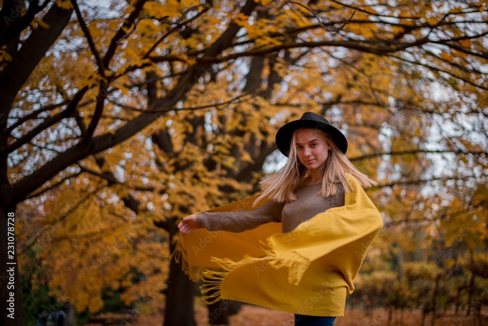 Beautiful blonde girl in a hat and yellow shawl dancing in autumn park full of yellow leaves