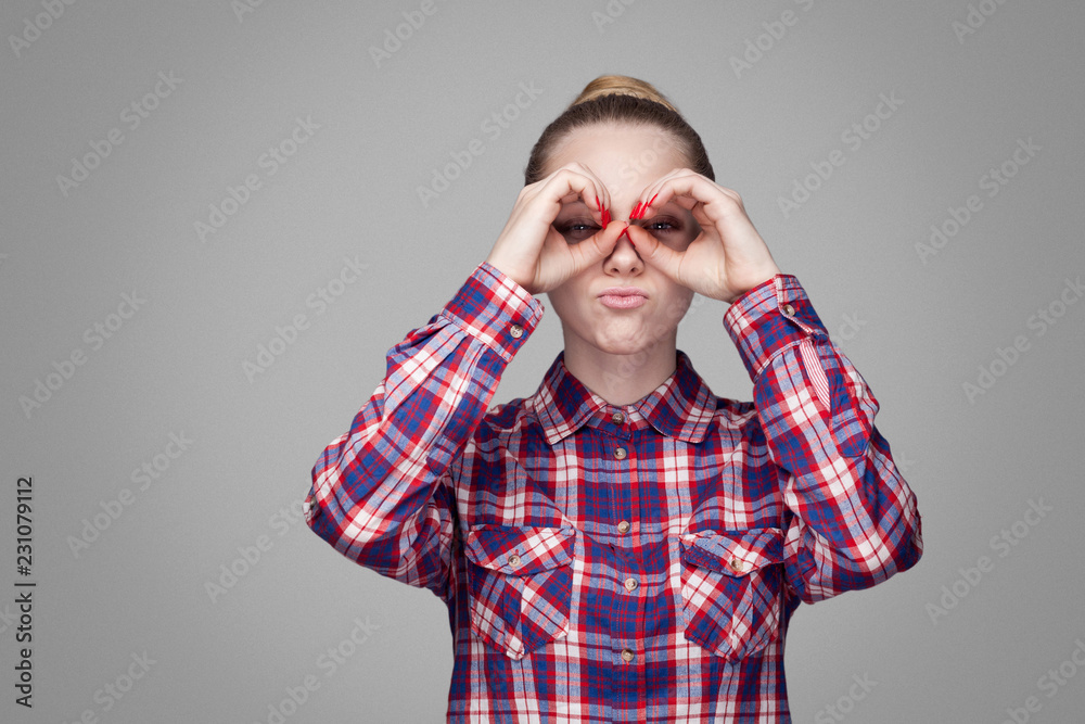 beautiful funny blonde girl in red, pink checkered shirt, collected bun hairstyle, makeup standing and looking at camera with binoculars gesture hands. indoor studio shot. isolated on gray background beautiful funny blonde girl in red, pink checkered shirt, collected bun hairstyle, makeup standing and looking at camera with binoculars gesture hands. indoor studio shot. isolated on gray background