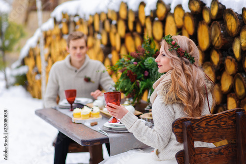 Groom kisses his bride on the temple. Newlyweds with bouquet sits on snow on the wooden background. Winter wedding photo