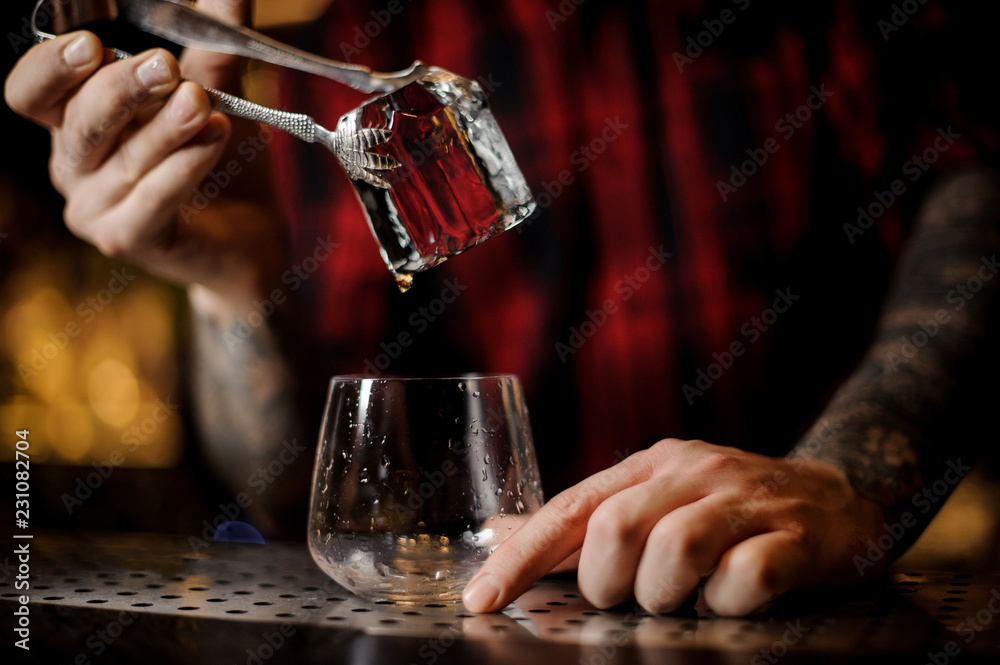 Bartender hand putting a big ice cube into a whiskey dof using tongs