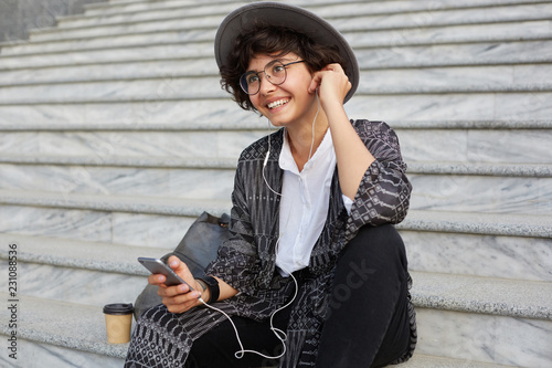 Portrait of young attractive cheerful woman sitting on the stairs, wears casula white shirt, cardigan and hat, listen a music via headphones and smiles. feels happy photo