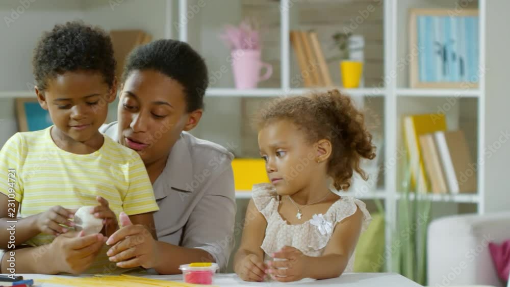 Tracking medium shot of cute little boy and girl making crafts with modeling clay, their mother and teacher talking to them and helping Tracking medium shot of cute little boy and girl making crafts with modeling clay, their mother and teacher talking to them and helping