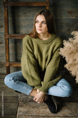 Sad brown haired dreamy model girl sitting cross-legged yoga style on wooden background floor. rustic studio in dark green knitted sweater and blue jeans. Fashion pose, arms on legs, looking away