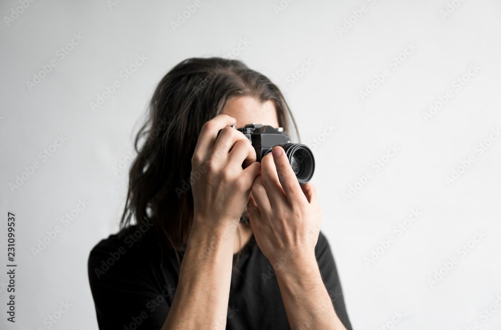 Handsome young bearded man with a long hair and in a black shirt holding vintage old-fashioned film camera on a white background and looking in camera viewfinder. Handsome young bearded man with a long hair and in a black shirt holding vintage old-fashioned film camera on a white background and looking in camera viewfinder.