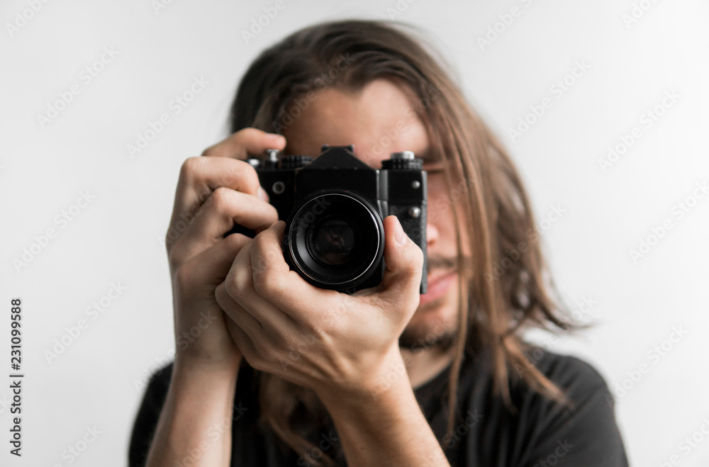 Handsome young bearded man with a long hair and in a black shirt holding vintage old-fashioned film camera on a white background and looking in camera viewfinder. Handsome young bearded man with a long hair and in a black shirt holding vintage old-fashioned film camera on a white background and looking in camera viewfinder.