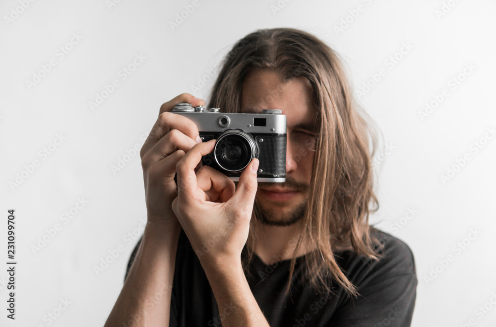 Handsome young bearded man with a long hair and in a black shirt holding vintage old-fashioned film camera on a white background and looking in camera viewfinder. Handsome young bearded man with a long hair and in a black shirt holding vintage old-fashioned film camera on a white background and looking in camera viewfinder.