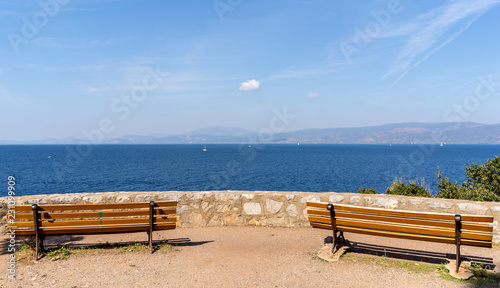 Two empty park benches on a stone overlook, with a view of a deep, blue sea and mountains in the distance.