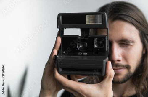 Handsome young bearded man with a long hair and in a black shirt holding vintage old-fashioned film camera on a white background and looking in camera viewfinder.