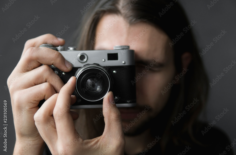 Handsome young bearded man with a long hair and in a black shirt holding vintage old-fashioned film camera on a black background and looking in camera viewfinder. Handsome young bearded man with a long hair and in a black shirt holding vintage old-fashioned film camera on a black background and looking in camera viewfinder.