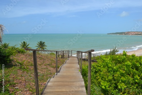 Caminho para o Paraiso feito de madeira artesanal, unindo a montanha à praia, com coqueiros e o mar azul ao fundo com um céu límpido