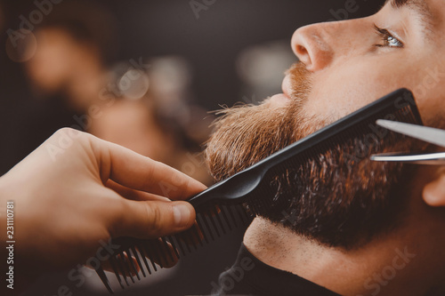 Hipster man sitting in armchair barber shop while hairdresser shaves beard with scissors. photo