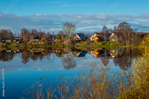 Fototapeta Naklejka Na Ścianę i Meble -  Blue River. Reflection of houses in the water. River bank and reflections in the water. Trees are reflected in the water. The village on the banks of the river.