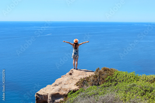 A girl stands on a cliff above the blue endless sea and enjoys the freedom and the view