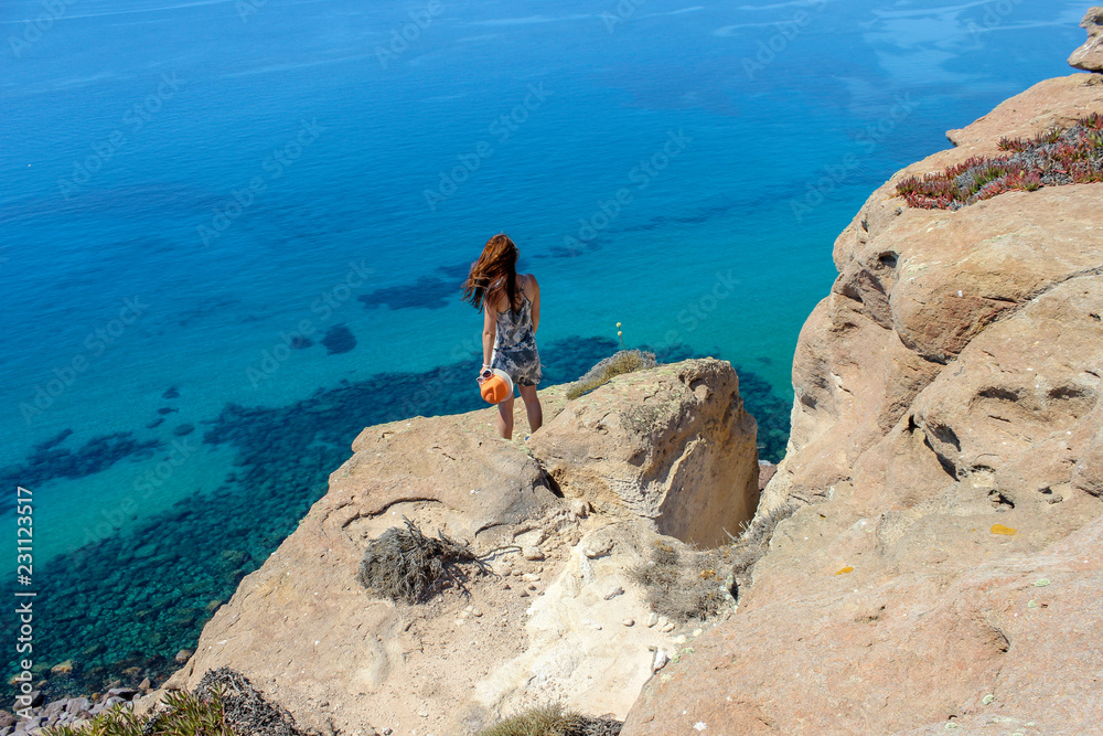 A girl stands on a cliff above the blue endless sea and enjoys the freedom and the view