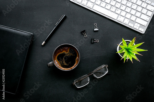 Black strict office desk, monochrome. Computer keyboard, expensive black notebook, glasses, coffee. Top view