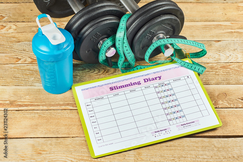 Fototapeta Naklejka Na Ścianę i Meble -  Healthy lifestyle concept. Mock up on workout and fitness dieting diary with copy space. Slimming diary sheet, measuring tape, blue shaker and dumbbells on a rustic wooden table