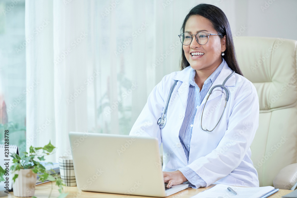 Cheerful adult Asian woman in doctor white coat sitting at desk with laptop smiling away while talking Cheerful adult Asian woman in doctor white coat sitting at desk with laptop smiling away while talking