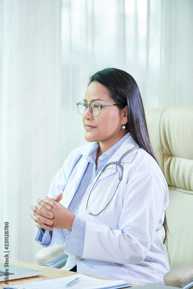 Smart professional Asian doctor in white coat sitting at table in office talking to patient Smart professional Asian doctor in white coat sitting at table in office talking to patient