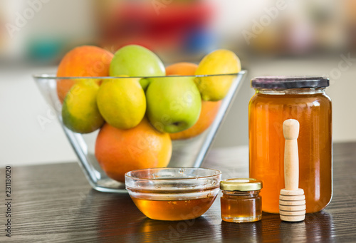 Big and small glass jar of honey with wooden sipper spoon and bowl of fruits on the background.