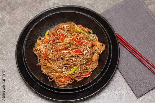 Fried noodles with vegetables, chicken and sesame in a black bowl. Gray background. Close up