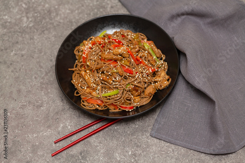 Fried noodles with vegetables, chicken and sesame in a black bowl. Gray background. Close up photo