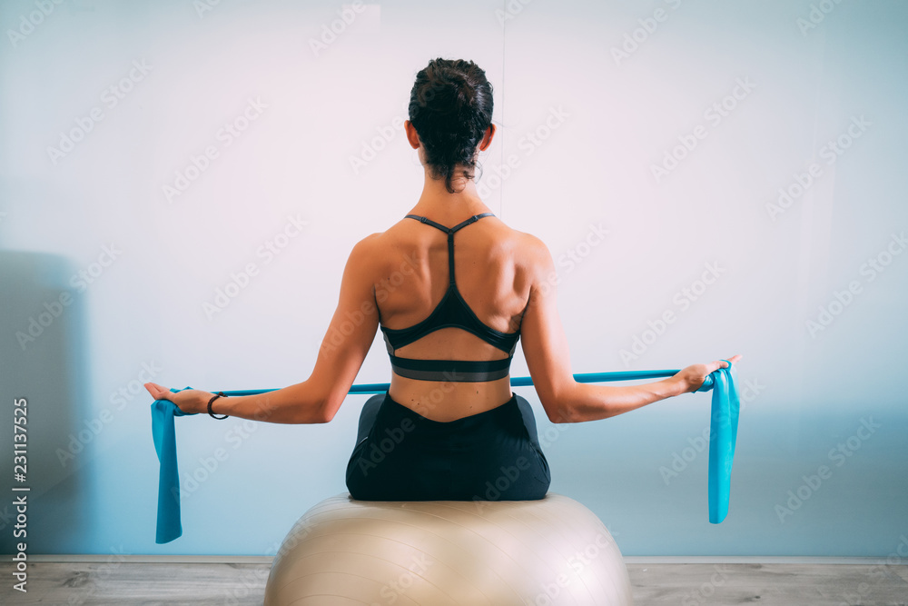 Young sporty attractive woman practicing pilates with resistance bands back sitting on fitness balls on white urban studio. Yoga and Pilates lifestyle healthy concept. - obrazy, fototapety, plakaty 