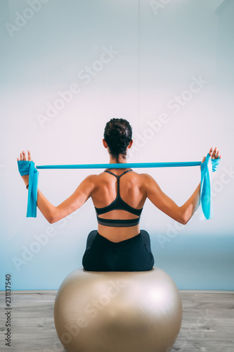 Фотография Young sporty attractive woman practicing pilates with resistance bands back sitting on fitness balls on white urban studio