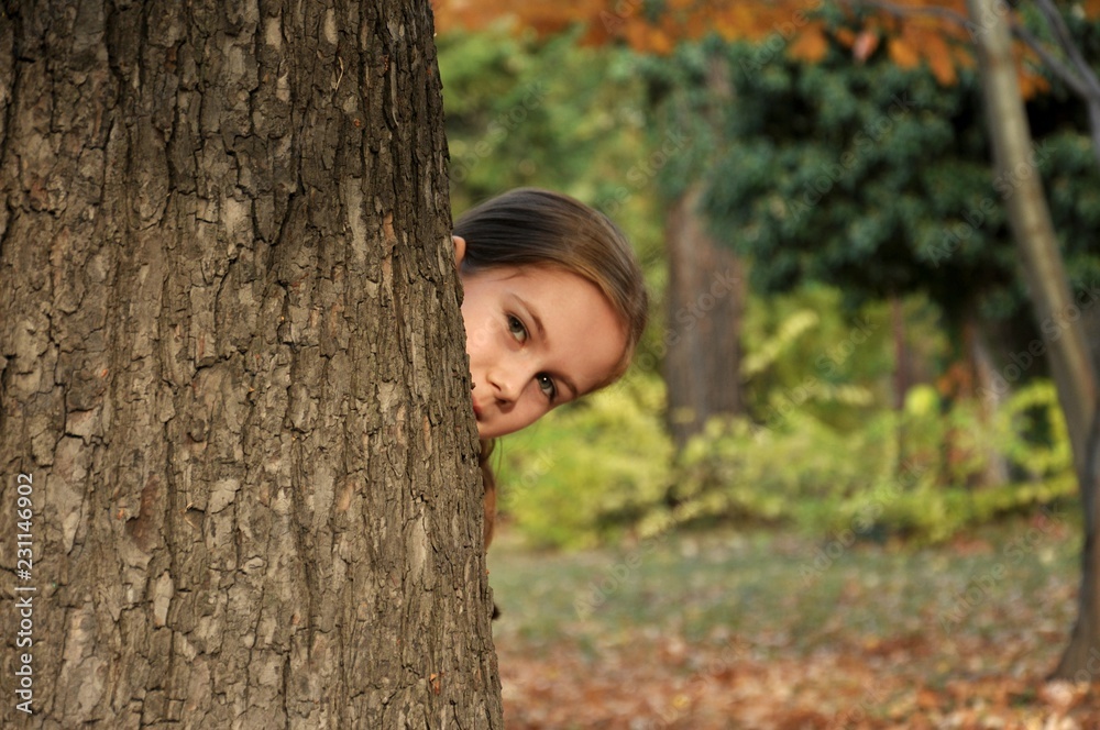 Caucasian 8-9 years old child, girl, happy in the park, smiling and hiding behind a big tree Caucasian 8-9 years old child, girl, happy in the park, smiling and hiding behind a big tree